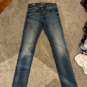 Slim Glenn jeans från Jack & Jones - Snygga blå jeans från Jack & Jones i modellen Slim Glenn. Jeansen har en slim passform, klassisk femficksdesign och snygga slitningar framtill. Tillverkade i mjukt denimtyg med lite stretch för extra komfort. Perfekta för en avslappnad och trendig look. Flärpen där bak har lossnat lite därför sänkt pris men löttt att du fst igen. Original pris runt 899