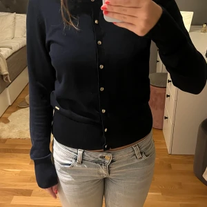 Mörkblå kofta från Holly & Whyte - Snygg mörkblå kofta från Holly & Whyte med rund hals och silvriga knappar framtill. Långärmad och i mjuk bomull, perfekt att slänga över ett linne eller t-shirt. Klassisk och enkel modell som funkar till allt.