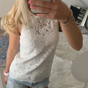 Vit spetsig ärmlös blus - Säljer en vit blus med spetsmönster och hög hals. Toppen är ärmlös och har en snygg, feminin look med detaljerad spets över hela plagget. Perfekt för dig som gillar romantisk stil och vill ha något luftigt och fräscht till sommaren.