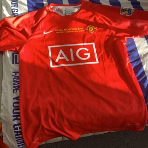 Manchester United Ronaldo 7 tröja - Säljer en röd Manchester United fotbollströja från Nike med AIG som sponsor, Premier League-märke på ärmen och tryck för Champions League-finalen 2008. På ryggen står Ronaldo och nummer 7. Tröjan är kortärmad och tillverkad i lätt, ventilerande material.