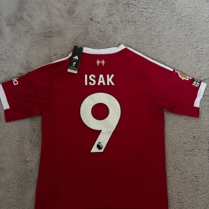 Liverpool FC Isak #9 Adidas tröja - Liverpool FC matchtröja från Adidas med Isak och nummer 9 på ryggen. Röd med vita detaljer, broderat LFC-märke, Premier League-logga och sponsortryck. Kortärmad och tillverkad i ventilerande polyester. Snygga vita ränder på axlarna och klassisk passform.