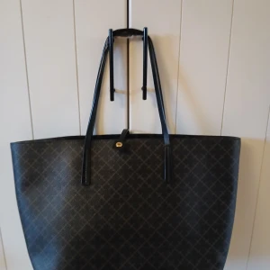  By Malene Birger - Stor och rymlig shoppingväska från By Malene Birger i mörkbrunt med ett snyggt rutigt mönster. Väskan har svarta handtag och är tillverkad i slitstarkt syntetmaterial. Perfekt för dig som gillar stilren design och vill ha plats för allt du behöver.