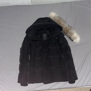 Svart dunjacka från Canada Goose wyndham med äkta päls - Säljer en svart dunjacka från Canada Goose med stor huva och snygg pälskant. Jackan har klassisk patch på ärmen och stängs med både dragkedja och knappar. Perfekt för kalla dagar och riktigt stilren look.