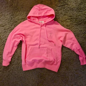 Rosa hoodie från AMI Paris - Säljer en snygg rosa hoodie från AMI Paris med huva och snörning. Tröjan har en broderad logga med hjärta på bröstet och en stor magficka. Tillverkad i mjuk bomull och har långa ärmar med ribbade muddar. Perfekt för dig som gillar streetwear vibes.