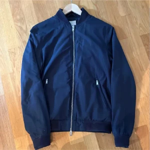 Marinblå bomberjacka från J.Lindeberg - Snygg marinblå bomberjacka från J.Lindeberg med klassisk passform. Jackan har dragkedja framtill, ribbade muddar och två fickor med dragkedja. Perfekt för dig som vill ha en stilren och enkel look.