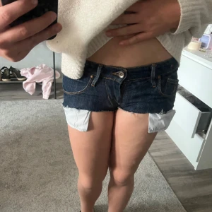 Mörkblå denimshorts med fransar - Säljer ett par mörkblå denimshorts med låg midja och råa fransade kanter. Shortsen har klassiska fickor fram och bak, samt synliga vita fickfoder som sticker ut under kanten. Perfekt för dig som gillar en edgy och avslappnad stil.(gjort själv)