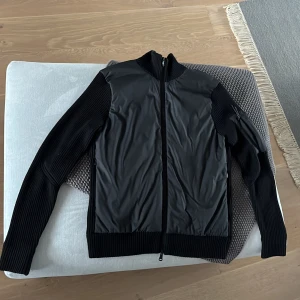 Svart hybridjacka med ribbade ärmar - Snygg svart jacka med glansig framsida och ribbstickade ärmar och rygg. Jackan har hel dragkedja framtill och hög krage. Perfekt för dig som gillar en stilren look med sportig touch.