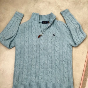 Ljusblå kabelstickad tröja Polo Ralph Lauren - Kabelstickad ljusblå tröja från Polo Ralph Lauren med halv dragkedja och broderad logga på bröstet. Tröjan har hög krage, långa ärmar och ribbade muddar. Materialmix av ull, akryl och nylon gör den både mjuk och varm.