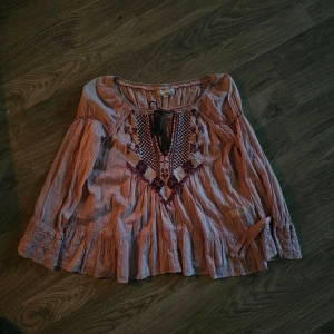 Odd Molly broderad blus i rosa - Broderad blus från Odd Molly i ljusrosa bomull med volangkant, långa ärmar och knytning vid halsen. Framsidan har broderier i vinrött och vitt, och baksidan har texten 'welcome inside my imagination lab' broderad. Perfekt för dig som gillar boho-stil. Står inte storlek men gissar S eller M