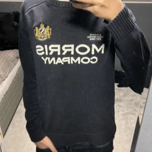 Mörkblå stickad tröja från Morris - Säljer en mörkblå stickad tröja från Morris Company med broderad logga och emblem på bröstet. Tröjan har rund hals, ribbade muddar och vit text framtill. Perfekt för dig som gillar klassisk och stilren look.