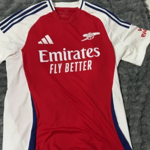 Arsenal röd och vit matchtröja Adidas - Säljer en Arsenal fotbollströja från Adidas i rött och vitt med blå detaljer. Tröjan har korta ärmar, klassiska Adidas-ränder på axlarna och klubbmärke samt Emirates-tryck på bröstet. Tillverkad i lätt och ventilerande material, perfekt för träning eller match.