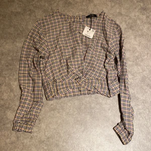Rutig croppad blus från Bershka XS - Säljer en croppad rutig blus från Bershka i storlek XS. Toppen har långa ärmar, twistad omlott-detalj framtill och ett mönster i svart, vit, gul och lila. Perfekt för dig som gillar trendiga och färgglada plagg med snygg passform.