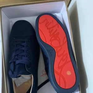 Mörkblå sneakers Christian Louboutin - Snygga mörkblå sneakers från Christian Louboutin med lyxig röd signatursula och rund tå. Skorna har snörning och är tillverkade i mocka med detaljer i skinn. Perfekt för dig som vill sticka ut med exklusiv stil och ikonisk design.