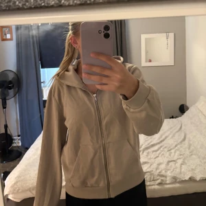 Hoodie - Säljer en beige hoodie med dragkedja framtill och huva. Tröjan har långa ärmar, ribbade muddar och två fickor framtill. Perfekt för en avslappnad och chill stil. Materialet känns mjukt och skönt mot huden.