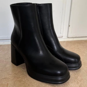 Svarta stövletter med klack, storlek 38 - Säljer ett par svarta stövletter i skinnimitation med chunky klack och dragkedja på sidan. Helt oanvända! 