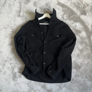 Svart overshirt från J.Lindeberg - Svart overshirt från J.Lindeberg i ull, med klassisk krage och knappar framtill. Två bröstfickor med lock och knapp, samt långa ärmar med manschettknappar. Perfekt för lager-på-lager och snygg till jeans eller chinos.