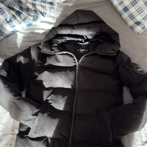 Svart pufferjacka Supply & Demand S - Svart pufferjacka från Supply & Demand med huva och dragkedja framtill. Jackan har en snygg badge på ärmen och breda, quiltade sektioner som ger en cool look. Perfekt för kalla dagar när du vill hålla stilen.