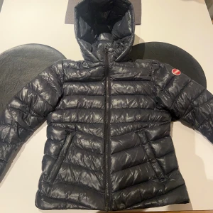 Svart Colmar pufferjacka med huva - Säljer en svart pufferjacka från Colmar med glansig finish och huva. Jackan har dragkedja framtill, två fickor med dragkedja och Colmar-logga på ärmen. Insidan är mönstrad med Colmar-tryck. Perfekt för kalla dagar och riktigt snygg streetstil.