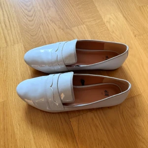 Vit/beigea lackloafers från H&M - Snygga vit-beiga loafers från H&M i storlek 37. Skorna har en glansig lackfinish, klassisk pennyloafers-design och spetsig tå. Perfekta för dig som gillar stilrena och trendiga skor med platt sula. Den har en liiiten defekt (bild 4) så därav sänker jag priset. 