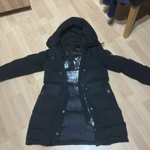 Svart lång pufferjacka Hollies - Svart lång pufferjacka från Hollies med huva och fuskpälsdetalj. Jackan har dragkedja, knappar och 3 fickor. En knapp har gått av men går att fixa innan den blir såld, kommer med päls ifall man vill köpa pälsen blir det en extra 300 eftersom pälsen är äkta. Köpte den för ungefär 2,5 år sen och den är i bra skick. Perfekt för vintern!