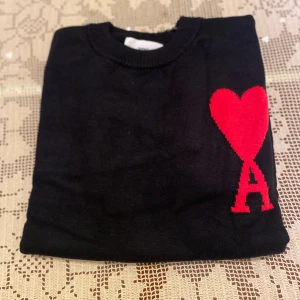 Svart stickad tröja från Ami Paris - Svart stickad tröja från Ami Paris med rund halsringning och ikonisk röd hjärtlogga med bokstaven A på bröstet. Tröjan har en klassisk passform och är perfekt för dig som gillar stilrena detaljer och streetstyle-vibe.