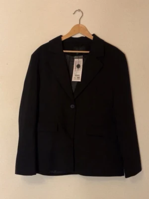 Svart kavaj - Klassisk svart kavaj med enkelknäppning och två fickor framtill. Rak passform och helfodrad insida. Perfekt till jeans eller kjol för en clean och stilren look. Materialet är mjukt och känns bekvämt mot huden. Helt ny, aldrig använd! Den är i storlek 42 men sitter som M/38