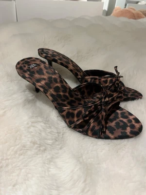 Leopardmönstrade sandaler från Zara - Säljer ett par sandaler från Zara i storlek 39 med leopardmönster. Skorna har låg klack, öppen tå och en snygg rosettdetalj framtill. Materialet känns mjukt och har en satinliknande finish. Perfekta för dig som vill sticka ut med ett djurmönster.