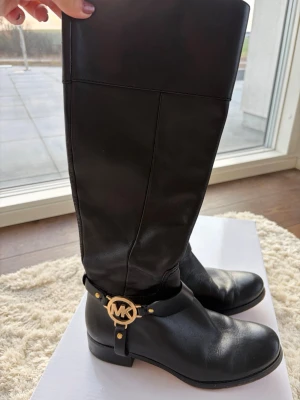 Svarta skinnstövlar Michael Kors - Snygga svarta stövlar från Michael Kors i äkta skinn med guldfärgad MK-detalj vid ankeln och dragkedja på insidan. Klassisk rund tå och låg klack gör dem perfekta till många outfits. Insidan är mjuk och sulan är i gummi.