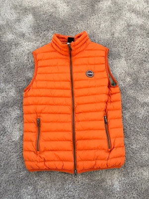 Colmar väst strlk 166 cm orange - Säljer en riktigt snygg orange dunväst från Colmar. Västen har quiltad design, hög krage och två fickor med dragkedja framtill. Perfekt att slänga över en hoodie eller tröja för extra värme. Klassisk Colmar-logga på bröstet och hel dragkedja framtill.