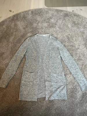 Ljusgrå kofta från Name It - En enkel och stilren ljusgrå kofta från Name It. Koftan har lång ärm, två fickor framtill och är tillverkad i ett mjukt och tunt material. 