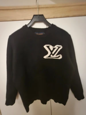Svart Lv tröja - Svart stickad tröja med en stor vit logga framtill. Tröjan har rund halsringning och långa ärmar. Perfekt för dig som gillar en enkel men ändå snygg look med en tydlig detalj på bröstet. Dock lite söndrig på sidan men märks inte av!