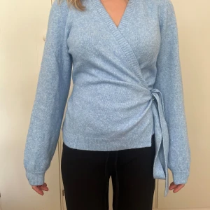 Ljusblå omlottkofta med knytband - Supersöt ljusblå omlottkofta med knyt i sidan och ribbade muddar. Tröjan har lång ärm och är stickad i mjukt material, perfekt för lager på lager under kyliga dagar. Enkel att styla till både jeans och kjol. Oanvänd, men tvättrådslappen på insidan är bortklippt.