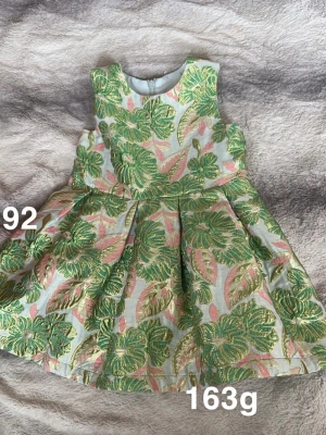 Blommig kortklänning i grönt och rosa - Superfin kortklänning med grönt och rosa blommönster på ljus botten. Klänningen har ärmlös design, rund hals och dragkedja bak. Materialet känns lyxigt och har en lätt glans, perfekt för speciella tillfällen.🌸 Fint använt skick. Storlek 92 