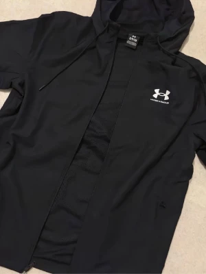 Svart vindjacka från Under Armour - Säljer en svart vindjacka från Under Armour med huva och dragsko. Jackan har dragkedja framtill och logga i vitt på bröstet. Tillverkad i lätt polyester som passar perfekt för träning eller när du vill ha en sportig look.