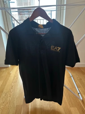 Svart piké från Emporio Armani EA7 - Säljer en svart pikétröja från Emporio Armani EA7 med guldfärgad logga på bröstet. Klassisk krage och knappar framtill. Tillverkad i mjuk bomull som känns skön mot huden. Perfekt för dig som vill ha en stilren och exklusiv look.