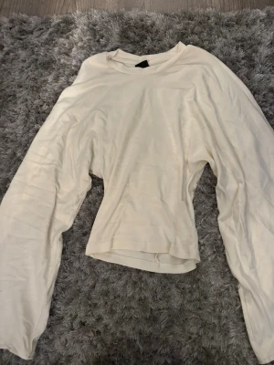 Vit långärmad topp Gina Tricot - En enkel och stilren vit långärmad topp från Gina Tricot i storlek XS. Toppen har rund halsringning och är gjord i ett mjukt, tunt material som känns skönt mot huden. Perfekt att matcha med jeans eller kjol för en clean look. Figurnära med stora ärmar. Aldrig använd.