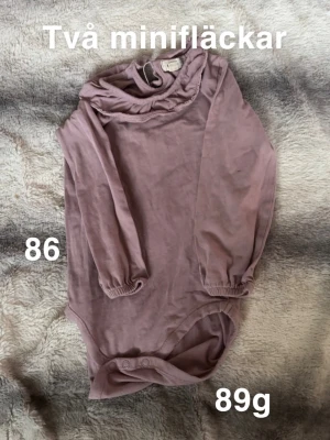 Lila långärmad body med volangkrage - Säljer en söt långärmad body i lila med volangdetalj vid kragen. Bodyn har knäppning vid grenen och resår vid ärmsluten för extra komfort. Perfekt för dig som gillar mjuka och stilrena plagg till de små.🌸 Två mini fläckar. Storlek 86