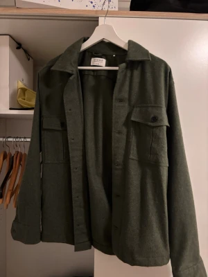 Olivgrön overshirt från Gabba - Säljer en stilren olivgrön overshirt från Gabba. Jackan har två stora bröstfickor med knappar, klassisk krage och knäppning framtill. Perfekt lager-på-lager-plagg med avslappnad passform och lång ärm. Materialet känns mjukt och har en ullig känsla.
