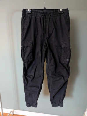 Svarta cargopants med snörning - Svarta cargopants från Smith & Soul med elastisk midja och snörning. Byxorna har flera praktiska fickor på sidorna och resår vid bensluten för en chill look. Tillverkade i bomull, perfekta för dig som gillar streetstyle och funktion.