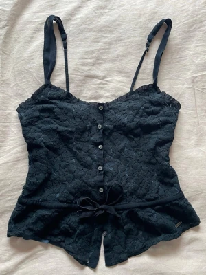 Marinblått spetslinne Hollister - Marinblått spettslinne från Hollister i storlek XS men skulle säga att den är mer S. Toppen har justerbara axelband, knappar framtill och är dekorerad med spets över hela ytan. Midjan har ett band med rosett och insidan är fodrad med mjukt tyg. 