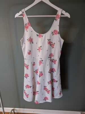 Vit blommig kortklänning med rosa rosor - Superfin vit kortklänning med rosa rosor och gröna blad. Klänningen är ärmlös med breda axelband och har en lätt utställd kjol. Perfekt för varma dagar och ger en söt och fräsch look. Materialet känns mjukt och bekvämt mot huden.