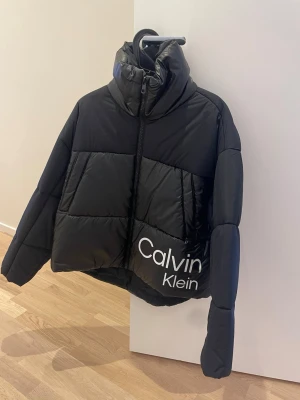 Svart pufferjacka Calvin Klein Jeans XS - Svart pufferjacka från Calvin Klein Jeans i storlek XS. Jackan har hög krage, stor vit logga på framsidan och dragkedja hela vägen. Den är quiltad och har en loose passform som ger en chill vibe. Perfekt för kalla dagar när du vill vara både varm och snygg.