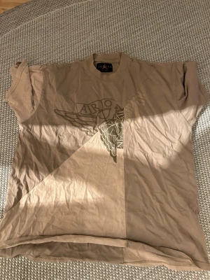 Beige Jordan x Cactus Jack t-shirt - Säljer en beige Jordan x Cactus Jack t-shirt med coolt vingtryck och basketboll på framsidan. Baksidan har broderad kaktus och texten 'Cactus Jack'. Oversized fit och mjuk bomull gör den riktigt skön. Perfekt för dig som gillar streetwear och snygga detaljer.