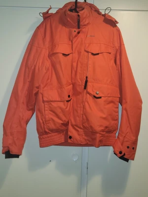 Orange vinterjacka från Quechua L - Säljer en orange vinterjacka från Quechua, perfekt för kalla dagar ner till -10°C. Jackan har flera stora fickor med knappar, huva och dragkedja framtill. Insidan är quiltad och svart, vilket ger extra värme. Märke: Decathlon/Quechua.