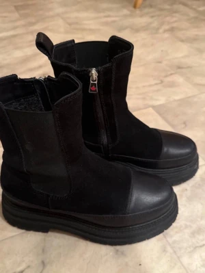 Svarta vinterskor från Canada Snow. Storlek 38 - Säljer ett par chunky svarta boots från Canada Snow med dragkedja på sidan och grov sula. Skorna är i en mix av mocka och skinn, har rund tå och platt sula. Perfekta för höst och vinter när du vill ha både stil och funktion. Endast använda ett fåtal gånger. Storlek 38