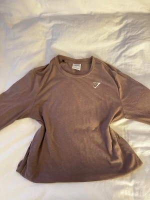 Brun långärmad t-shirt från Gymshark - ALDRIG använd!!! Säljer en brun långärmad t-shirt från Gymshark i storlek S. T-shirten har en enkel design med rund hals och Gymshark-logga tryckt på bröstet. Materialet är mjuk bomull som känns skönt mot huden. Perfekt för dig som gillar stilrena träningskläder.