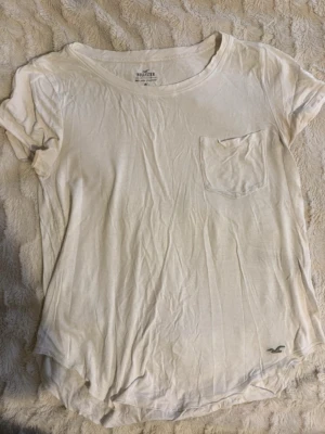 Vit t-shirt från Hollister - Enkel vit t-shirt från Hollister med rund halsringning och bröstficka. T-shirten har korta ärmar och en liten Hollister-logga nere, lite genomskinlig och har en avslappnad passform.