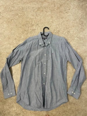 Blå skjorta från Tommy Hilfiger M - Snygg blå skjorta från Tommy Hilfiger i storlek M. Klassisk button-down krage, vita knappar och diskret logga på bröstet. Skjortan är långärmad och har en normal passform. Perfekt till jeans eller chinos för en clean look.