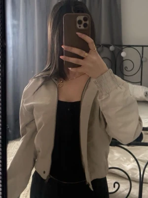 Beige bomberjacka med dragkedja - Säljer en beige bomberjacka med dragkedja framtill och resår vid ärmslut och midja. Jackan har en clean och enkel look, perfekt till lager-på-lager. Lätt croppad passform och långärmad modell. Passar dig som gillar stilrena och neutrala plagg.