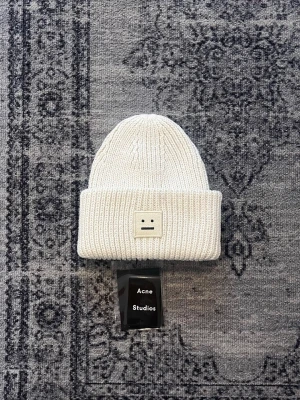 Vit stickad mössa från Acne Studios - Vit ribbstickad mössa från Acne Studios med det klassiska face-logotypen framtill. Mössan är i mjukt och varmt material, perfekt för kyliga dagar. Enkel och stilren design som passar till det mesta.